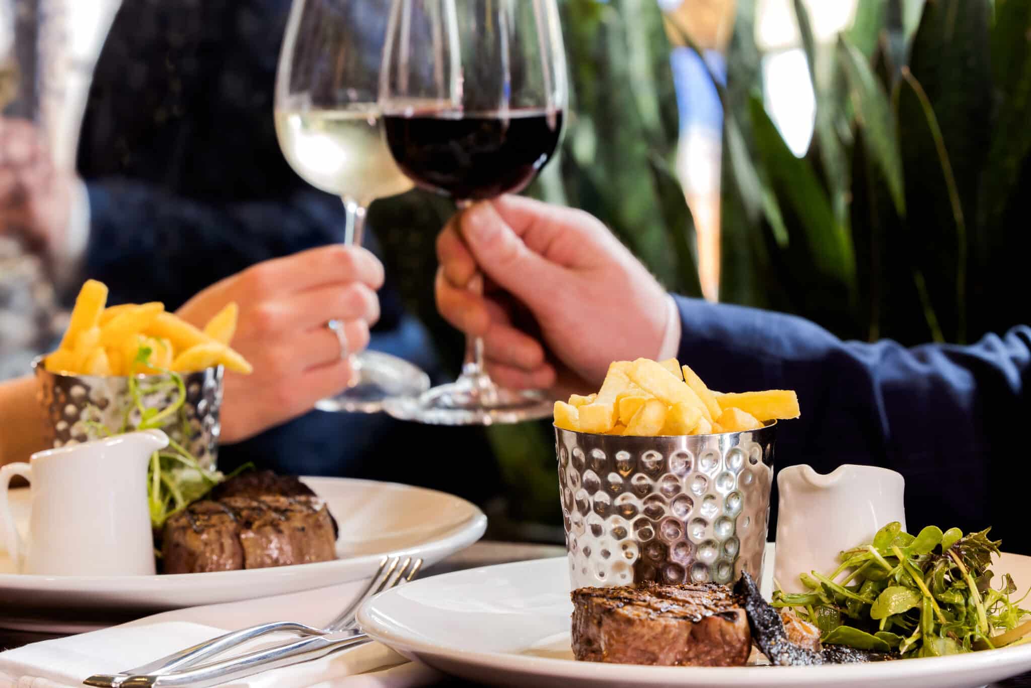 Two people clinking wine glasses over a delicious steak dish, enjoying Black Friday at The Brasserie